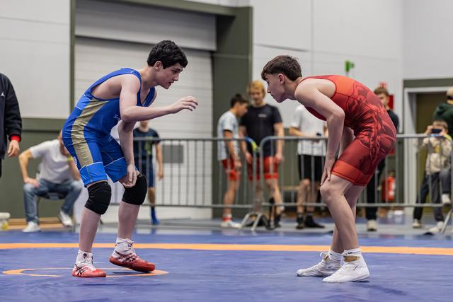 Two teen wrestlers in blue and red singlets crouch low, locked in intense eye contact before their match on a blue mat.