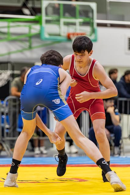 Two teen wrestlers in red and blue singlets square off on a yellow mat, the red wrestler focused and alert.