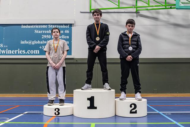 Three young boys stand on a winner's podium, wearing medals, in a sports hall. The first-place boy looks subdued.
