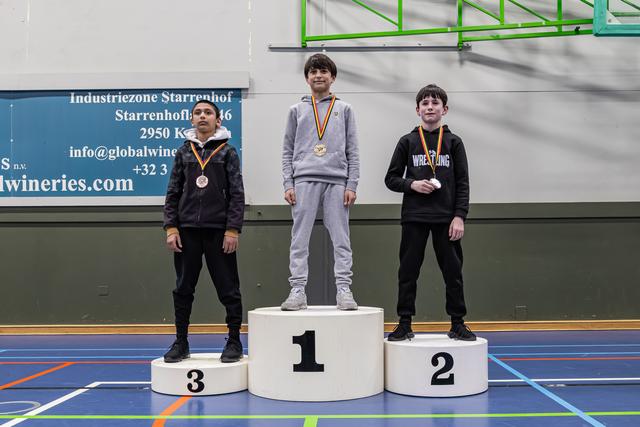 Three young wrestlers on a podium; gold medalist stands tall in center, flanked by bronze and silver winners, all wearing medals.