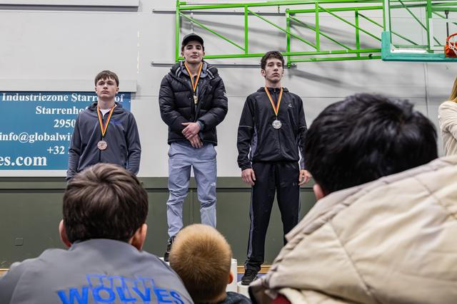 Three teenage wrestlers stand on a podium wearing medals, expressions serious and composed, in an indoor gymnasium.