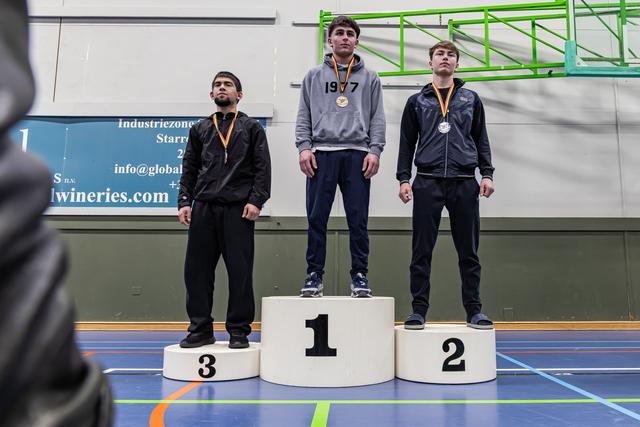 Three young wrestlers stand on a podium, wearing medals. The gold medalist stands tallest, center, looking composed.