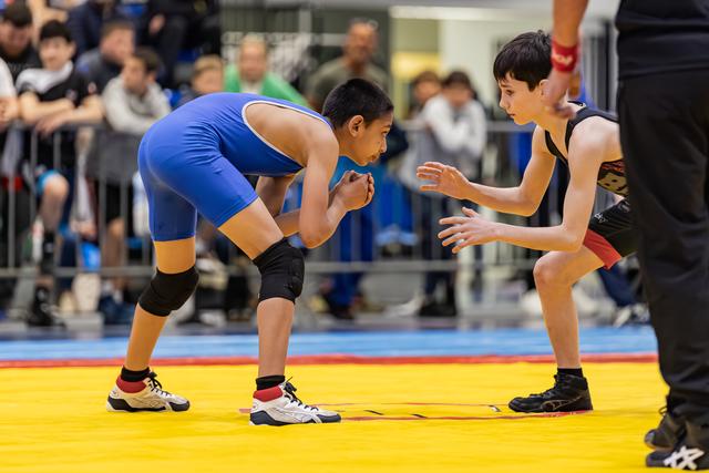 Two young wrestlers in blue and black singlets face off on a yellow mat, focused and tense, crowd watching behind.
