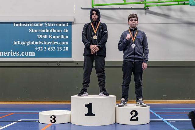 Two young boys stand on a winner's podium; the gold medalist looks composed while the silver medalist proudly displays his medal.
