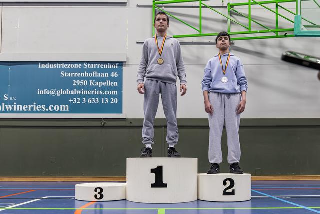 Two young athletes stand on podiums 1 and 2, wearing medals, expressions serious and composed, in an indoor sports hall.
