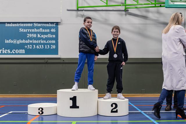 Two boys shake hands on a winner's podium, both smiling with medals around their necks in an indoor sports hall.