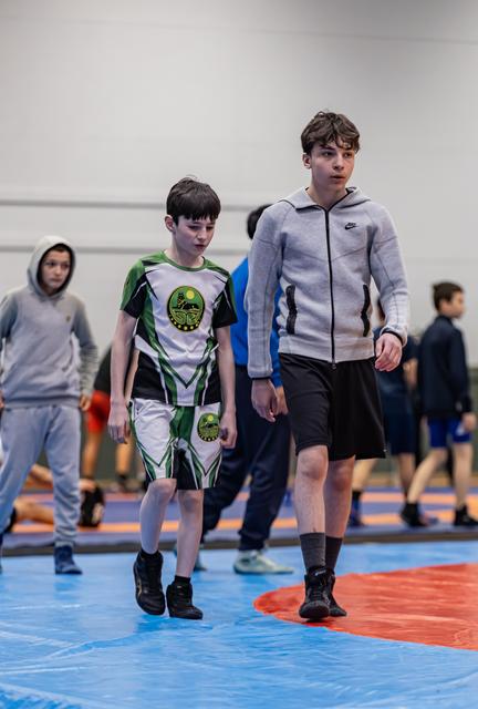 Two young wrestlers walk across the mat, the smaller boy in a green uniform looks downcast, the taller in a Nike hoodie walks beside him.