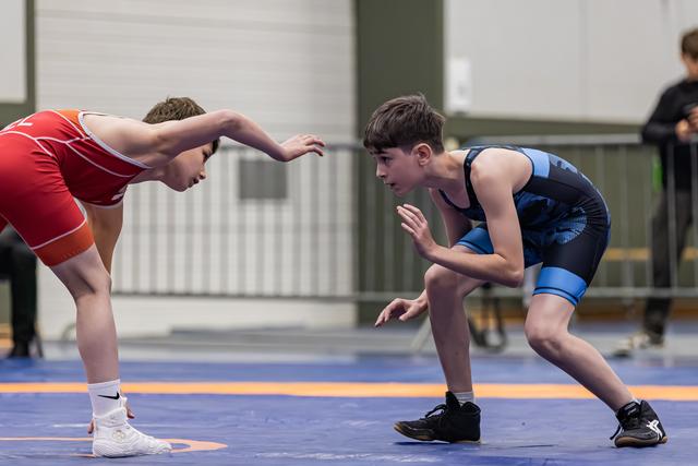Two young wrestlers in red and blue singlets face off in a crouched stance on the blue mat, focused and ready.