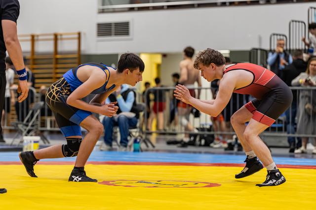 Two teenage wrestlers crouch in tense standoff on yellow mat, hands raised, focused expressions in a gymnasium.
