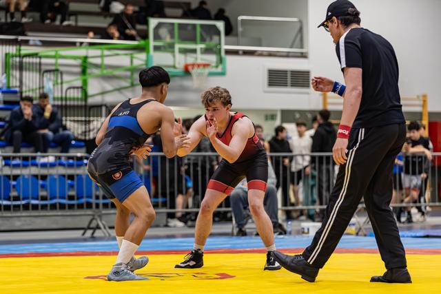 Two teen wrestlers face off in tense stance on yellow mat, referee watching closely in indoor sports hall.