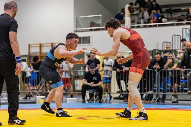 Two wrestlers in navy and red singlets face off in a tense stance, watched by a crowd at an indoor sports hall.