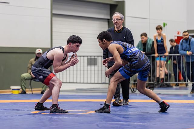 Two young wrestlers face off in intense stances on a blue mat, with a referee watching closely behind them.