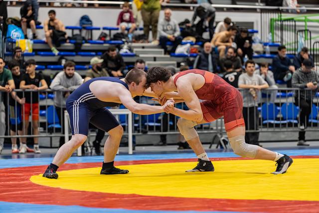 Two wrestlers in navy and red singlets lock hands in an intense face-off on a colorful mat before a packed crowd.