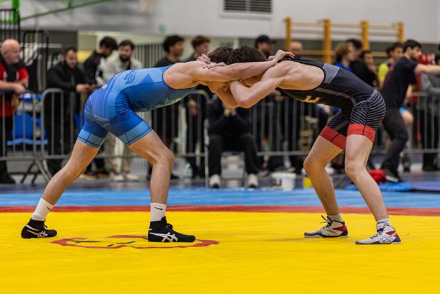 Two wrestlers lock heads in a tie-up stance on a yellow mat, crowds watching in the background.