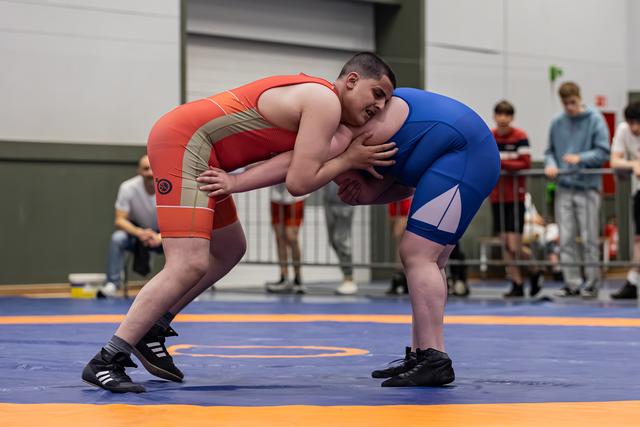 Two young wrestlers lock arms in an intense clinch on a blue mat, straining forward with determined effort.