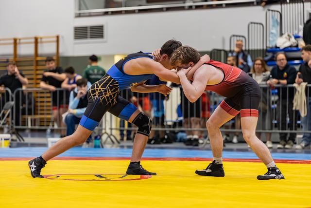 Two young male wrestlers grip each other in a standing clinch, both low and intense on a yellow competition mat.