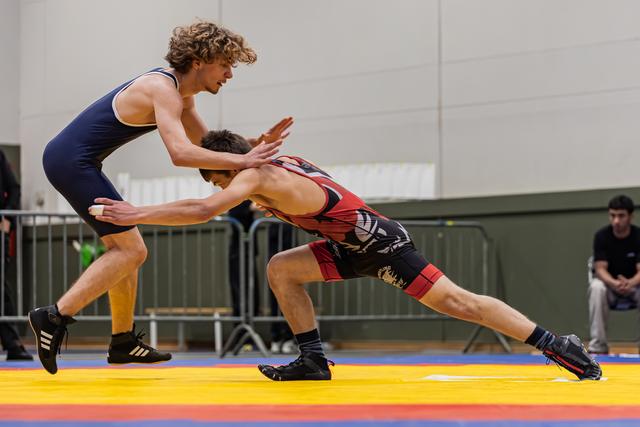 Curly-haired wrestler in blue singlet defensively pushes back opponent lunging low in red, on colorful mat.