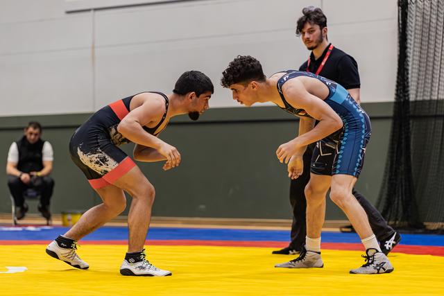 Two young wrestlers in a tense standoff, leaning toward each other on a yellow mat, referee watching closely behind.