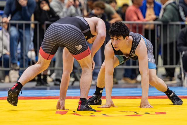 Two young wrestlers crouch in a tense standoff on a yellow mat, the facing athlete showing focused concentration.