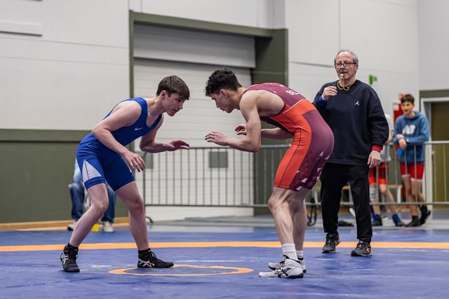 Two young wrestlers square off in intense stance on blue mat, referee watches closely in background.