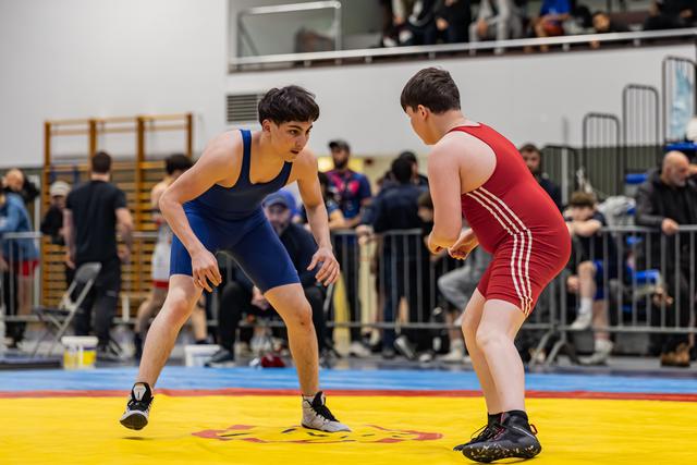 Two young wrestlers in blue and red singlets crouch in tense standoff on colorful mat, crowd watching behind barriers.