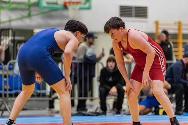 Two young wrestlers in blue and red singlets crouch low, locked in intense eye contact before engaging on the mat.