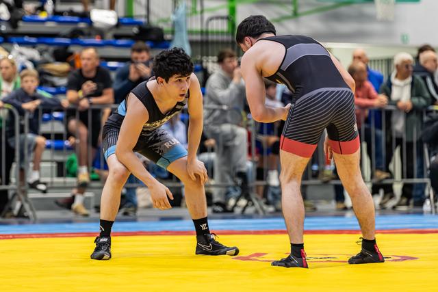Two young wrestlers in black singlets face off in a crouched, tense stance on a yellow mat before a crowd.