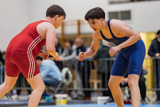 Two young wrestlers in red and blue singlets square off, crouched in tense ready positions on an indoor mat.