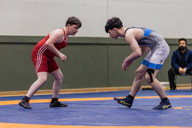 Two young wrestlers in red and grey singlets square off intensely on a blue competition mat, referee watching behind.