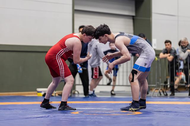 Two young wrestlers crouch face-to-face in a tense standoff on a blue mat, spectators watching in the background.