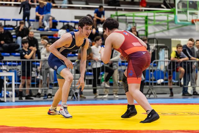 Teen wrestler in blue singlet squares off against opponent in red, both crouched in defensive stance on yellow mat.
