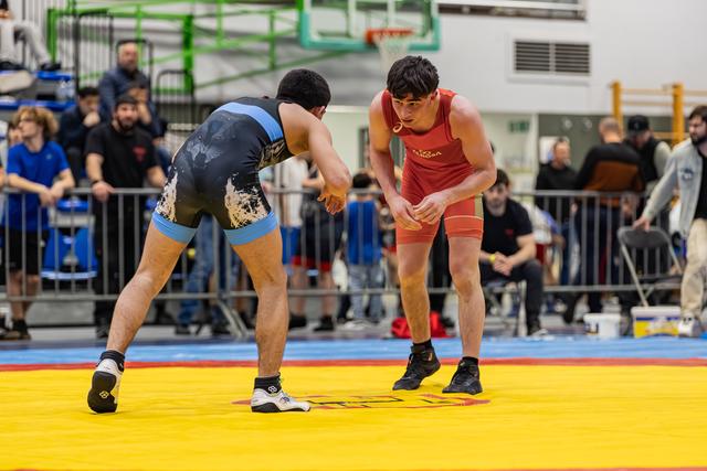 Two wrestlers in blue and red singlets crouch in tense standoff on a yellow mat, crowd watching behind barriers.