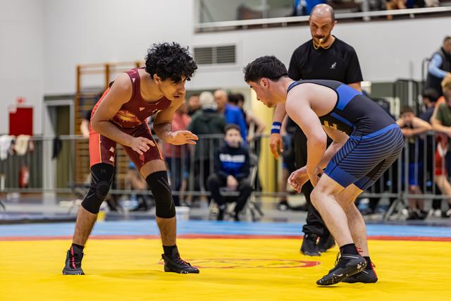 Two young wrestlers in red and blue singlets square off in a tense standoff, referee watching closely behind them.
