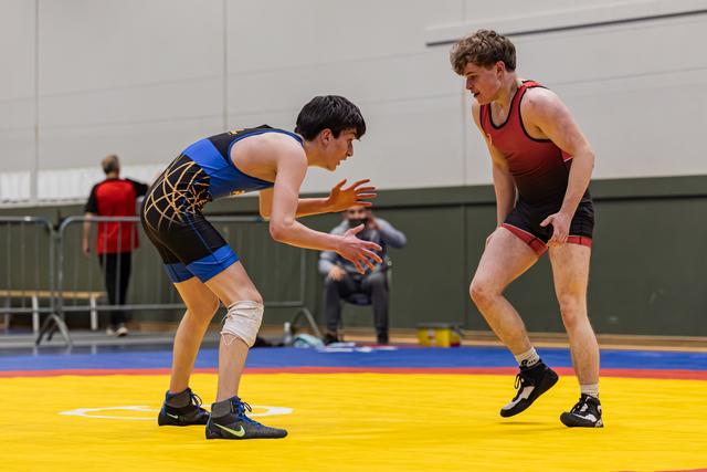 Two young wrestlers in blue and red singlets square off in a crouched stance, focused and ready to engage on a yellow mat.