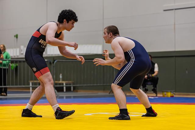 Two male wrestlers in fighting stance, circling each other intensely on a yellow mat in an indoor sports hall.