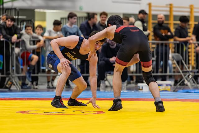 Two young wrestlers in a tense standoff, crouched low on a yellow mat, spectators watching closely in the background.