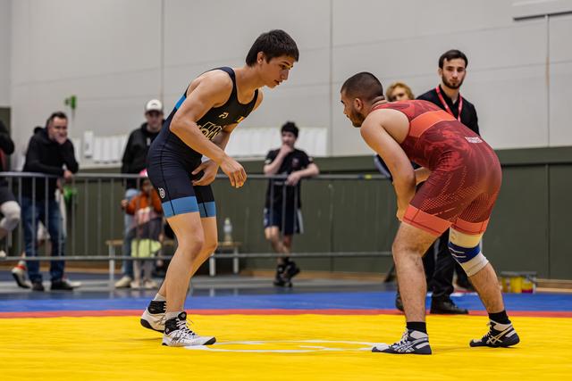Two wrestlers in blue and red singlets square off in a tense standoff on a yellow competition mat, crowd watching behind.