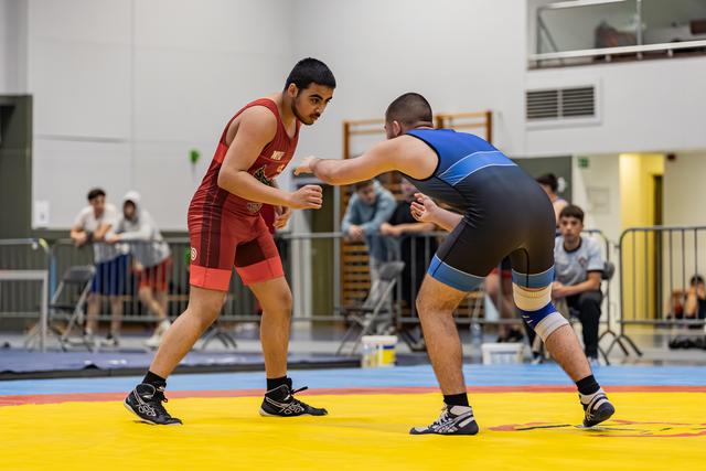 Two wrestlers in red and blue singlets square off on a yellow mat in an indoor sports hall, crowds watching.