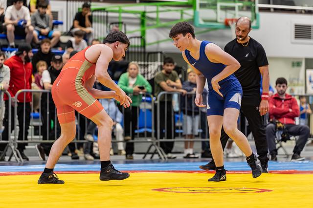 Two young wrestlers in orange and blue singlets crouch in a tense standoff on a yellow competition mat, referee watching closely.