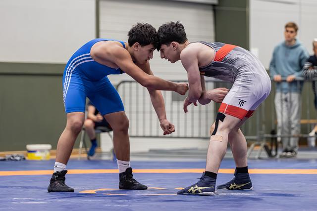 Two young wrestlers in blue and gray singlets lock up forehead-to-forehead in an intense grappling standoff on a blue mat.