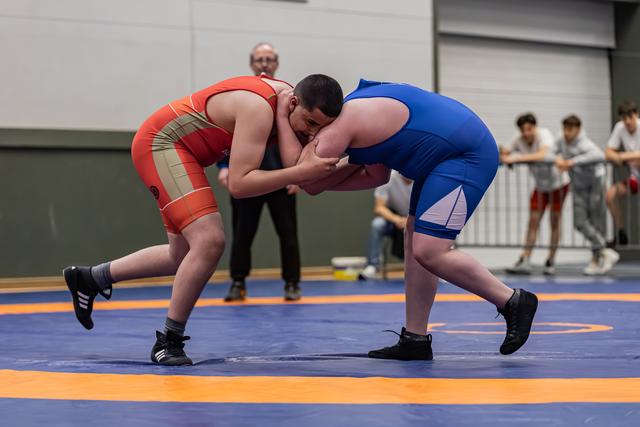 Two heavyweight wrestlers locked in a tight clinch on a blue mat, referee watching closely behind them.