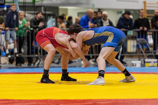 Two young wrestlers in red and blue singlets locked in a clinch, both bent low with intense focus on a competition mat.