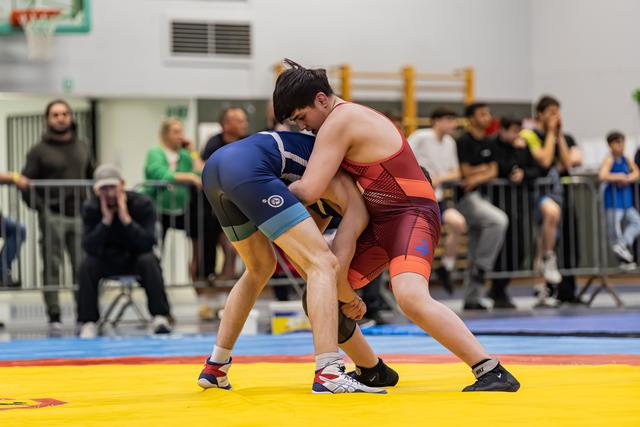 Two young wrestlers locked in a grapple on yellow mats, blue vs red singlet, crowd watching intently behind barriers.