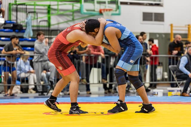 Two wrestlers locked in a clinch on a yellow mat, both hunched forward, straining for control in a busy gymnasium.