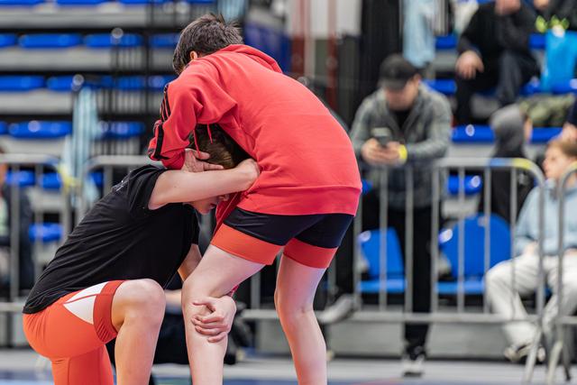 Two young wrestlers locked in a tight clinch on the mat, both leaning forward with intense focus during an indoor event.