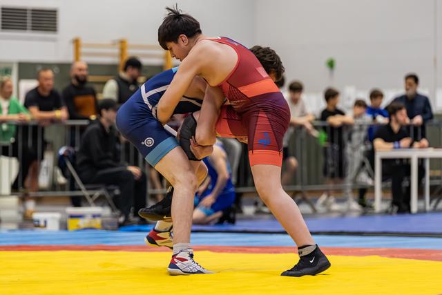 Two young wrestlers in blue and red singlets locked in a powerful clinch on a yellow mat, crowd watching behind.