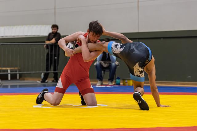 Young wrestler in red singlet on one knee, gripping opponent in dark blue singlet in intense clinch on yellow mat.