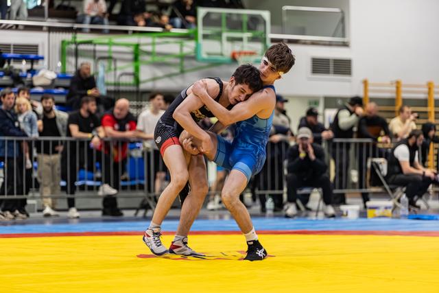 Two young wrestlers locked in an intense clinch on a yellow mat, crowd watching in a packed indoor sports hall.