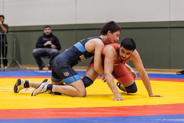 Young wrestler in blue singlet controls opponent in red from behind, both straining intensely on a colorful mat.