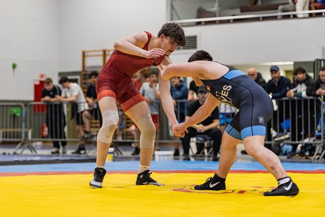 Two young male wrestlers lock grips in a tense standoff on a yellow mat, spectators watching from behind barriers.